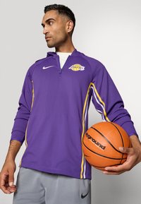 Lila Nike Sportpullover mit halbem Reißverschluss, ausgestattet mit gelben Streifen und einem Lakers-Logo. Dazu sind graue Shorts sichtbar, der Spieler hält einen Basketball.