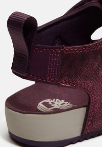 Gros plan sur le talon compensé bordeaux d'une sandale avec une bride texturée, un onglet de traction et une semelle beige arborant un logo d'arbre blanc.