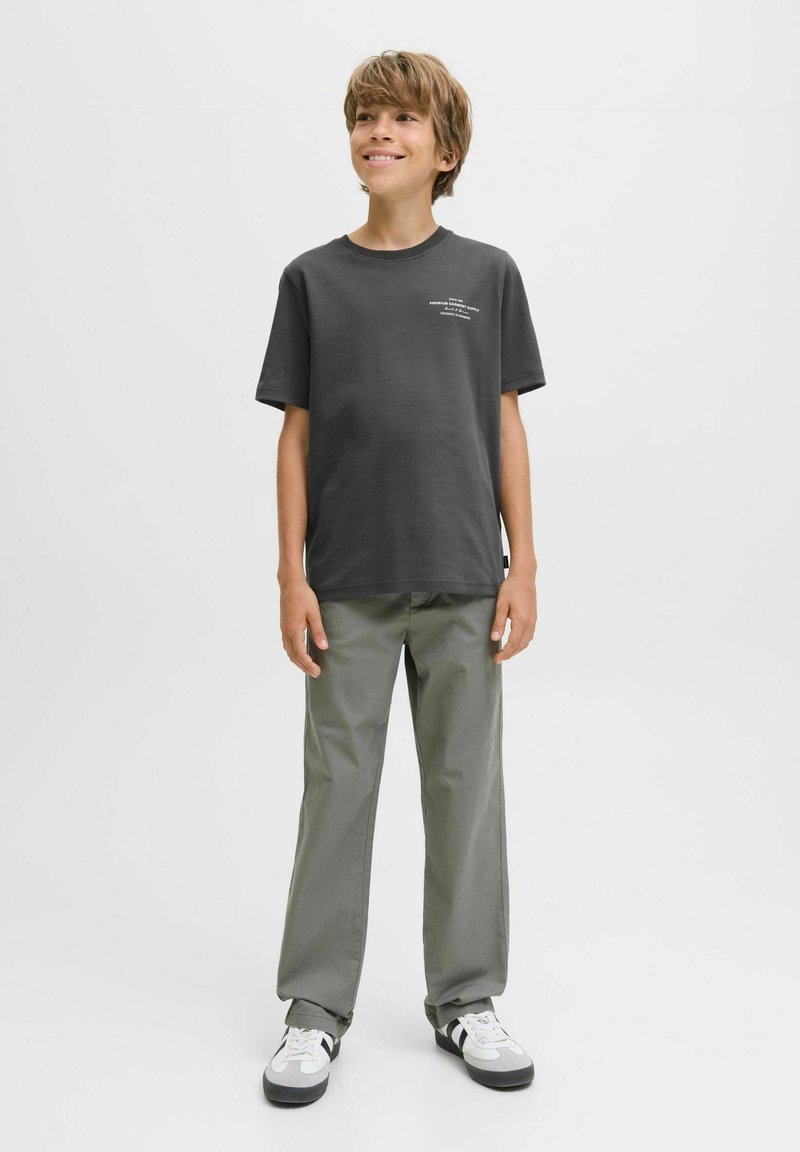 A smiling boy stands confidently, wearing a dark gray t-shirt and olive-green pants, with white sneakers against a light background.