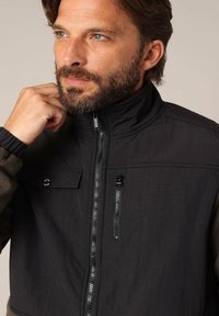 Zwarte en olijfgroene jas met ritssluiting, hoge kraag en voorzak. Beschikt over een textuurstof en een aansluitend ontwerp.