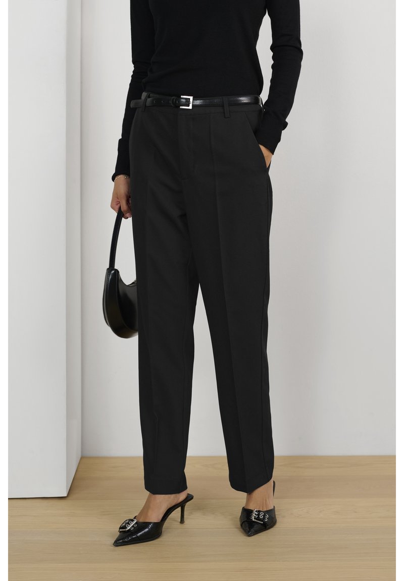Femme portant un pantalon noir taille haute sur mesure avec une ceinture noire, un haut noir à manches longues, des talons noirs à bout pointu, et tenant un sac à main noir.