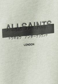 T-shirt di tessuto grigio chiaro con la scritta "ALLSAINTS" in ricamo nero, una striscia orizzontale nera e "LONDON" sotto.
