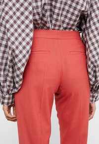 Rode op maat gemaakte broek met een gladde textuur, voorzien van een hoge taille, twee achterzakken en een plooidetail. Gecombineerd met een geruite blouse.
