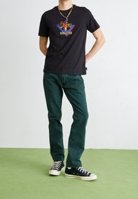 Person trägt ein schwarzes T-Shirt mit Grafik, grüne Jeans, schwarze High-Top-Sneaker und eine goldene Kette, steht auf einem grünen Boden vor einer weißen Wand.