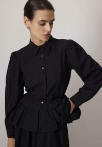 Žena s tmavými vlasy v černé strukturované košili na knoflíky s nafouknutými dlouhými rukávy a uvázaným pasem, dívající se doleva.