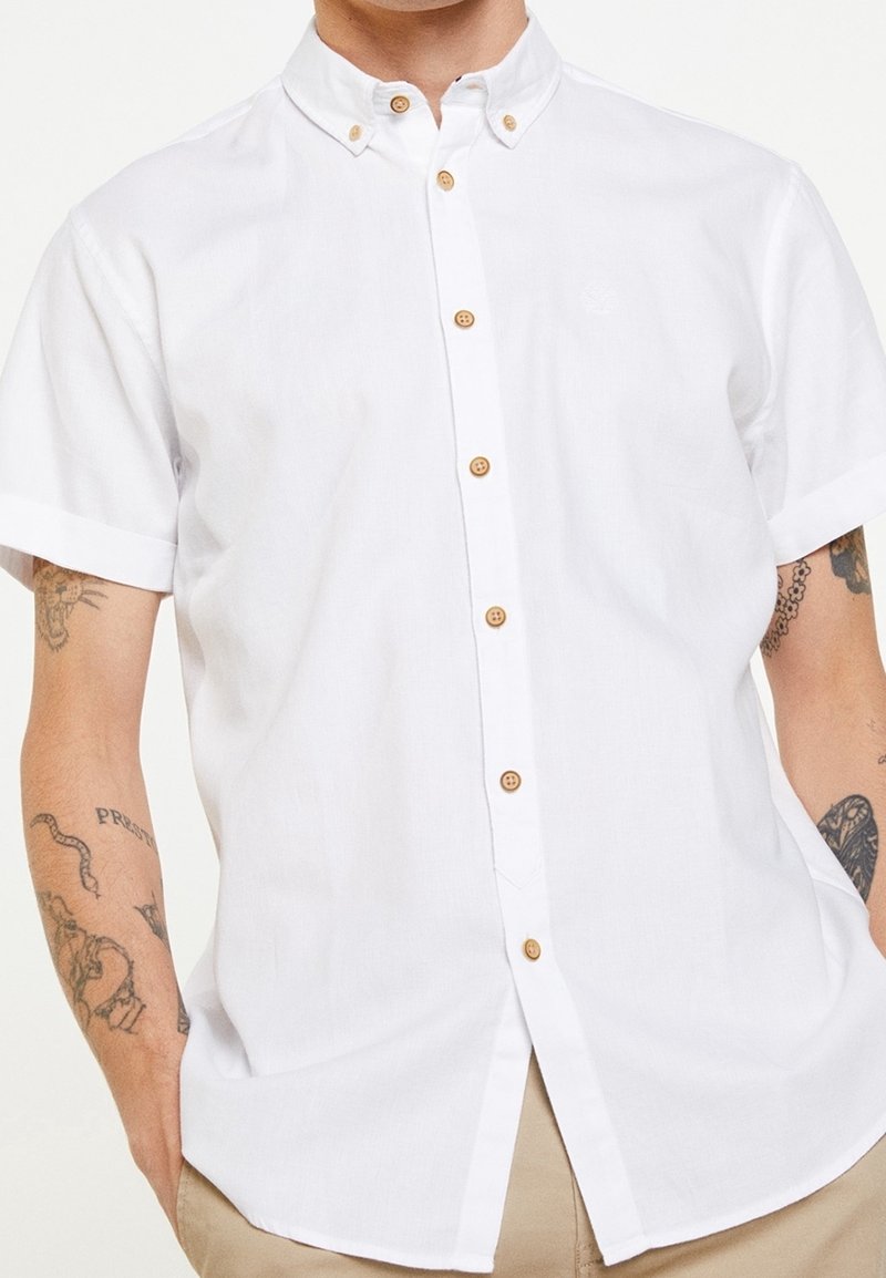 Chemise blanche à manches courtes en tissu léger, dotée de boutons en bois et d'un col classique, avec une coupe décontractée.