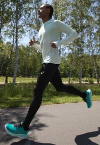 Chaussures de sport en bleu sarcelle avec des accents noirs, associées à un haut à manches longues vert menthe clair et à des leggings noirs, portées par une personne courant en plein air.