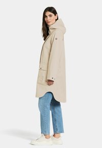 Beige waterdichte jas met een capuchon, sluiting met knopen aan de voorkant, een rond afgewerkte onderkant en twee zijzakken, gedragen boven lichtblauwe recht-gesneden jeans en witte sneakers.