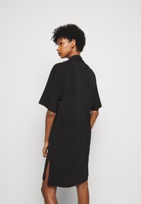 Svart kortärmad klänning med krage, sprund bak, och slät textur. Knälång design med rak silhuett.