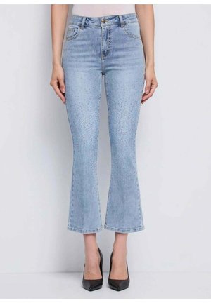 Donna che indossa jeans a zampa di elefante a vita alta e corta di colore azzurro chiaro con dettagli sottili di strass e décolleté nere con punta appuntita, in piedi su uno sfondo bianco.