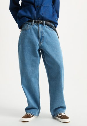 Person trägt locker sitzende hellblaue Jeans mit schwarzem Gürtel, dunkelblauem Hemd und braunen und weißen Sneakern und steht auf weißem Hintergrund.