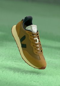 Sneaker marrone con materiali in rete e suede, caratterizzato da un logo bianco e nero, intersuola crema e suola in gomma rossa.