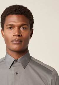 Szürke, gombos ing strukturált gallérral és texturált anyaggal, egyetlen gombbal a nyaknál és kontrasztos, átlós részletekkel.