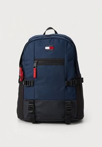 Námořnicky modrý batoh s hladkou tkaninou, černým dnem, červeným jezdcem zipu, nastavitelnými popruhy a přední kapsou na zip se vzorem v podobě diamantů.