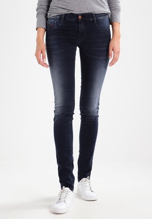 Personne portant un jean skinny bleu foncé, une chemise grise à manches longues et des baskets blanches, debout devant un fond blanc.