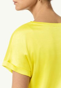 Krótki rękaw, żółta bluzka o gładkiej fakturze, szerokim dekolcie i złożonych mankietach. Tył jest delikatnie wykrojony z minimalnym detalem szwów.