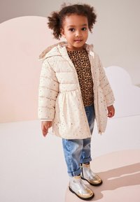 Beige pufferjakke med guld stjernet mønster, parret med en leopardprintet top og blå denimjeans. Sølv ankelstøvler med guld accenter.