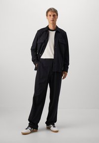 Uomo in piedi che indossa una giacca nera e pantaloni con una maglietta a righe bianche e sneakers bianche con strisce nere su sfondo chiaro.