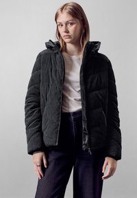 Giacca puffer in corduroy nera con chiusura a zip, superficie testurizzata e cappuccio. Indossata sopra una camicia bianca e jeans scuri.
