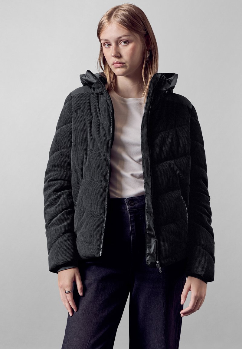 Giacca puffer in corduroy nera con chiusura a zip, superficie testurizzata e cappuccio. Indossata sopra una camicia bianca e jeans scuri.