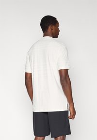 Koszulka sportowa o krótkich rękawach w kremowym kolorze z fakturowanym wzorem na plecach, zestawiona z ciemnymi szortami.