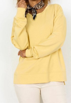 Persoon met een geel shirt met lange mouwen, licht beige broek en een bruine neksjaal met luipaardprint, poserend met één arm over de borst.