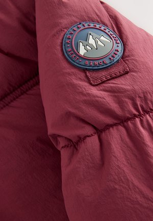 Mouw van een bordeauxrode donsjas met een rubberen patch met bergen en tekst. Soepele textuur met een naaddetail en nylon stof.