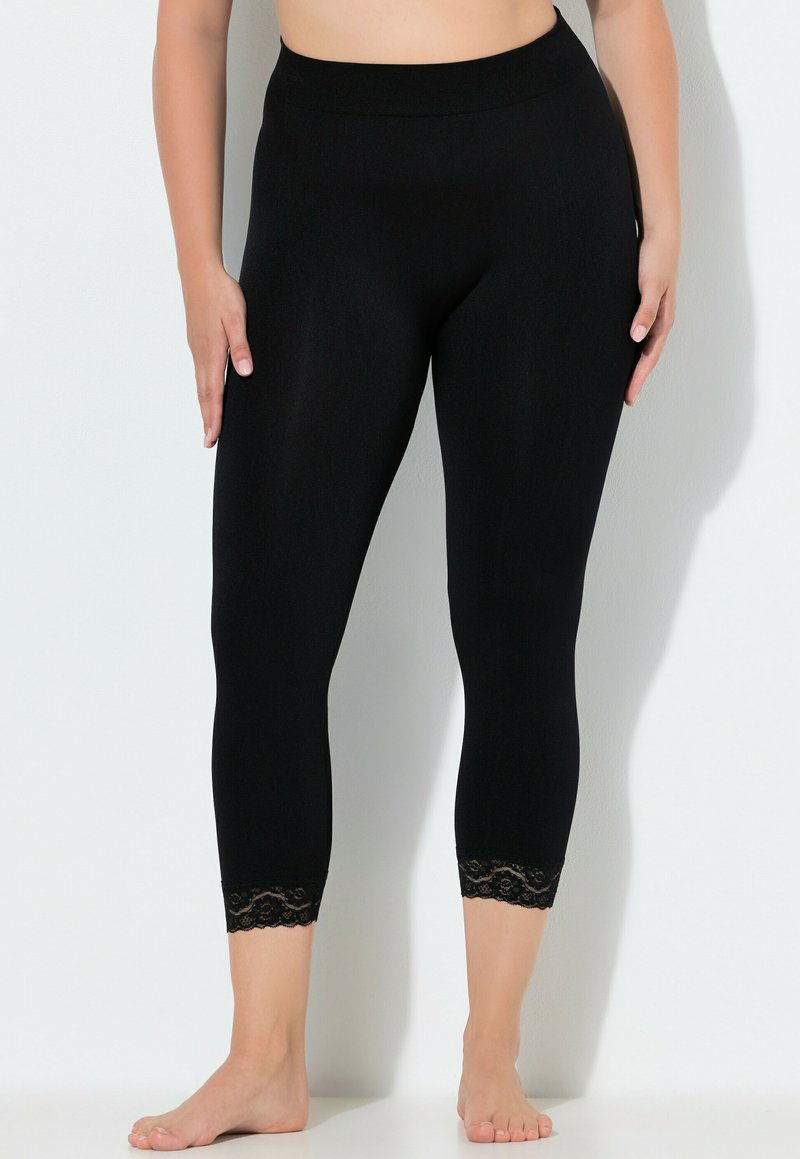 Mujer con leggings cortos negros de talle alto con encaje en los tobillos, de pie descalza contra una pared blanca.