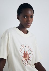 Cream-colored cotton t-shirt featuring a sun graphic in orange and maroon with the text "SLOW DOWN" styled around it. Relaxed fit, short sleeves.