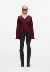 Pullover oversize bordeaux a scollo a V con maniche lunghe, abbinato a pantaloni neri aderenti e lucidi e stivali neri in pelle al ginocchio.