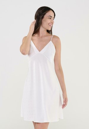 Mujer con cabello largo y oscuro, vestida con un vestido blanco sin mangas y tirantes finos, sonriendo y tocándose el cabello sobre un fondo claro simple.