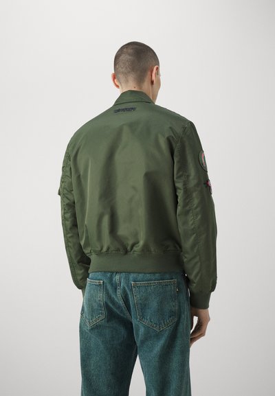 Uomo che indossa una giacca bomber verde con toppe sulle maniche e jeans blu sbiaditi, in piedi contro uno sfondo chiaro uniforme, visto da dietro.