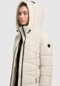 Kvadrični krem puffer jakna s visokim ovratnikom, velikim kapuljačom s vezicama, prednjim patentnim zatvaračem i okruglim crnim logotipom na rukavu.