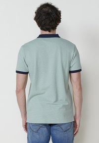 Zelené textured polo tričko s námořnickým límcem a akcenty na rukávech, s jednoduchým designem, zobrazené ze zadní strany, kombinované s džínovými kalhotami.