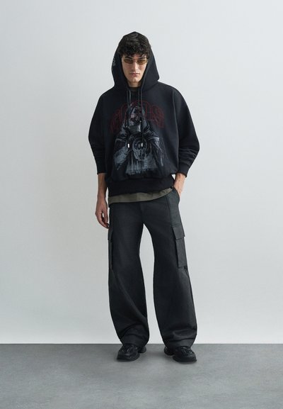 Hanorac negru cu imprimeu grafic, cămașă gri în straturi dedesubt, pantaloni cargo închiși la culoare și adidași negri, având o croială relaxată, supradimensionată.