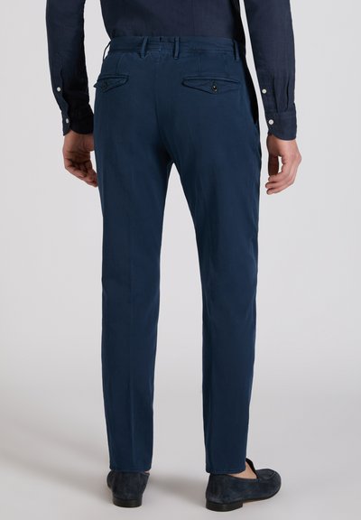 Man wearing navy blue slim-fit trousers and navy blue loafers, standing with hands relaxed by sides against plain background.
