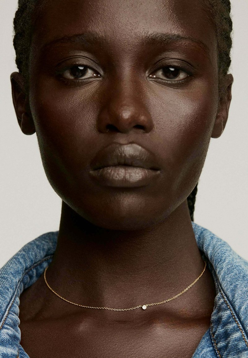 Collar de cadena de oro con un pequeño detalle de cuentas, llevado sobre piel oscura, complementado por el cuello de una chaqueta de mezclilla. Textura suave y diseño simple.