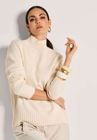 Frau in cremefarbenem Rollkragenpullover und Hose, mit großen Creolen und Chunky-Armbändern, posiert vor einem schlichten hellen Hintergrund.