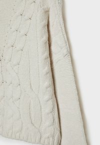 Tissu tricoté de couleur crème avec des motifs torsadés et côtelés, montrant la texture et le détail de la manche sur un fond uni.