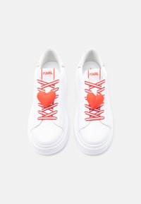 Λευκά αθλητικά παπούτσια με κόκκινες και λευκές κορδόνια, κόκκινα διακοσμητικά σε σχήμα καρδιάς στην κορυφή και λογότυπο "KARL" στη γλώσσα κάθε παπουτσιού.