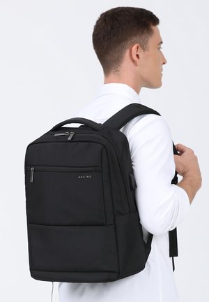 Sac à dos en tissu noir avec plusieurs compartiments zippés, de forme rectangulaire, avec des bretelles rembourrées et une poignée supérieure. Logo sur le panneau avant.