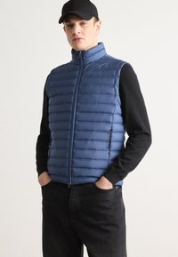 Chaleco acolchado azul con acolchado horizontal, cuello alto, cierre de cremallera y detalles en negro. Llevado sobre una camiseta de manga larga negra y vaqueros.