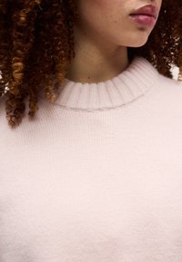 Κοντινό πλάνο ενός ατόμου με καστανά σγουρά μαλλιά που φοράει ένα ανοιχτό ροζ ριμπ πουλόβερ με στρογγυλή λαιμόκοψη, εμφανίζοντας τα χείλη, τον λαιμό και το πάνω μέρος του στήθους.