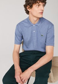 Blå melerad pikétröja med mjuk textur, utrustad med en tre-knapps placket och en grön broderad logotyp på vänster bröst.