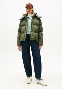 Veste matelassée olive verte avec un design matelassé, col montant et boutons-pression. Associée à une chemise rayée vert clair et un jean foncé.