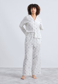 Biała piżama składająca się z koszuli zapinanej na guziki z kieszonką oraz spodni z sznurkiem, oba zdobione małymi niebieskimi kwiatowymi wzorami. Lekki materiał.