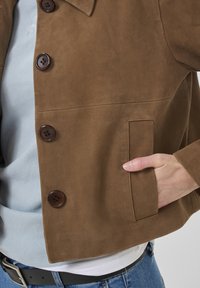 Brown suede jacket featuring a collar, six large buttons, a side pocket, and a smooth texture, layered over a light blue shirt.