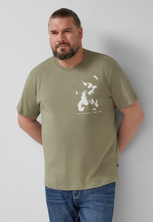 Man met baard en lang haar dat naar achteren is gebonden, draagt een olijfgroen T-shirt met een witte abstracte print en een blauwe spijkerbroek, staand tegen een effen muur.