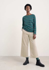 Moteris stovi vilkėdama žalią ir juodą dryžuotą ilgomis rankovėmis marškinį, šviesiai rudas plačias kelnes, žalias kojines ir juodas apdaras iki kulkšnies ant vienspalvio fono.