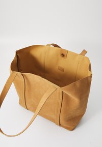 Sac fourre-tout en daim tan avec une texture douce, ouverture sur le dessus, deux poignées de épaule et une fermeture à bouton. L'intérieur possède un doublure lisse assortie.